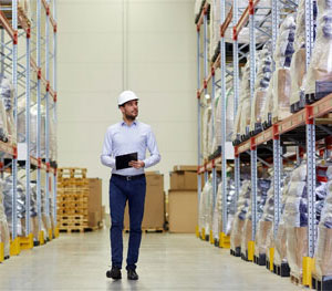 man with hard hat and clipboard walking through warehouse aisle