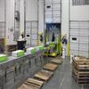 warehouse dock area with pallets on the floor and a worker standing near conveyor loaded with boxes