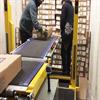 two men standing in front of a stack of boxes and placing boxes on conveyor